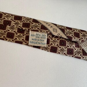 May include: A brown and beige patterned necktie featuring a repeating geometric square design. A white fabric label is stitched to the back, displaying the text 100% SETA-SILK SOIE-SEIDE MADE IN ITALY FABRIQUE EN ITALIE and a partial brand name.