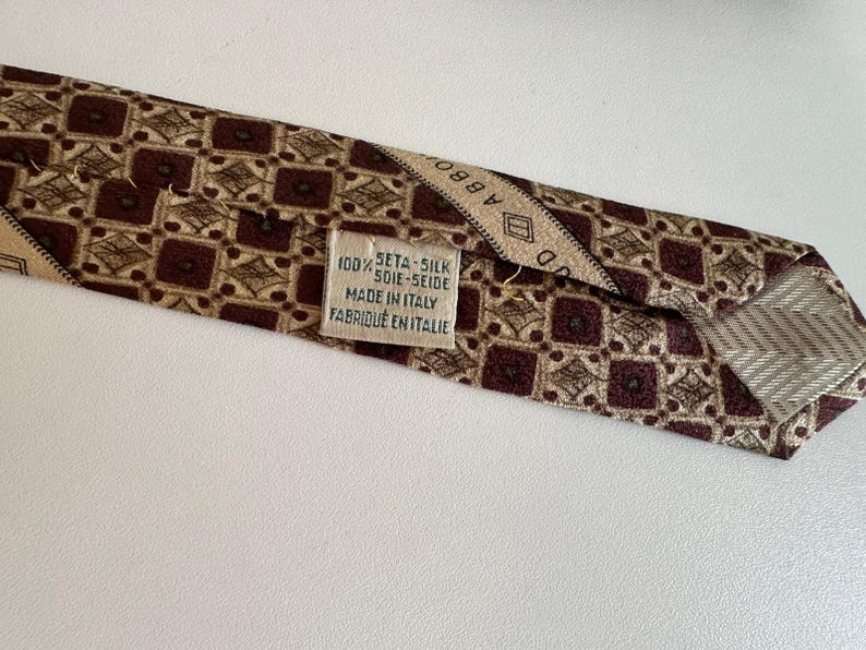 May include: A brown and beige patterned necktie featuring a repeating geometric square design. A white fabric label is stitched to the back, displaying the text 100% SETA-SILK SOIE-SEIDE MADE IN ITALY FABRIQUE EN ITALIE and a partial brand name.