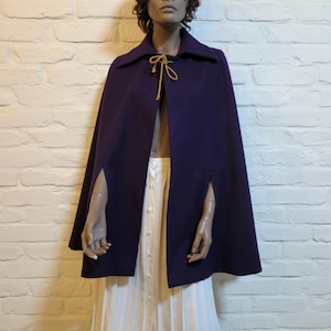 May include: A dark purple cape with a collar and arm slits. The cape has a gold-colored tie at the neckline. The mannequin is wearing a white dress underneath the cape. The background is a white brick wall.
