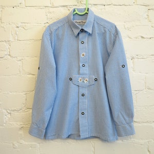 May include: A light blue and white gingham button-down shirt featuring a collar, long sleeves with roll-up tabs, and a front pocket flap. The shirt is decorated with embroidered white daisy motifs and includes the text "Ois & Toi" on the label.
