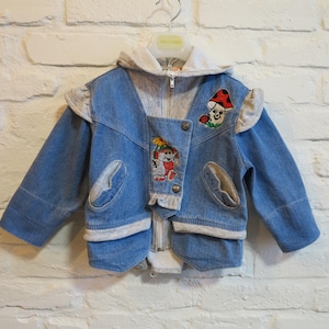 May include: A light blue denim jacket with a gray hood and accents. The jacket features embroidered cartoon characters and a mushroom design. It has a zipper closure and button details, perfect for children's wear.