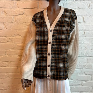 Puede incluir: Cárdigan abotonado con un estampado de cuadros en marrón, negro y blanco. Las mangas son de color beige liso y el cárdigan tiene un escote en V. La prenda está hecha de un material suave y texturizado.