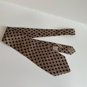 May include: A necktie featuring a repeating geometric pattern of dark brown squares and light beige diamond shapes. The tie has a pointed end and a visible white fabric label stitched onto the back side near the center, displaying printed text.