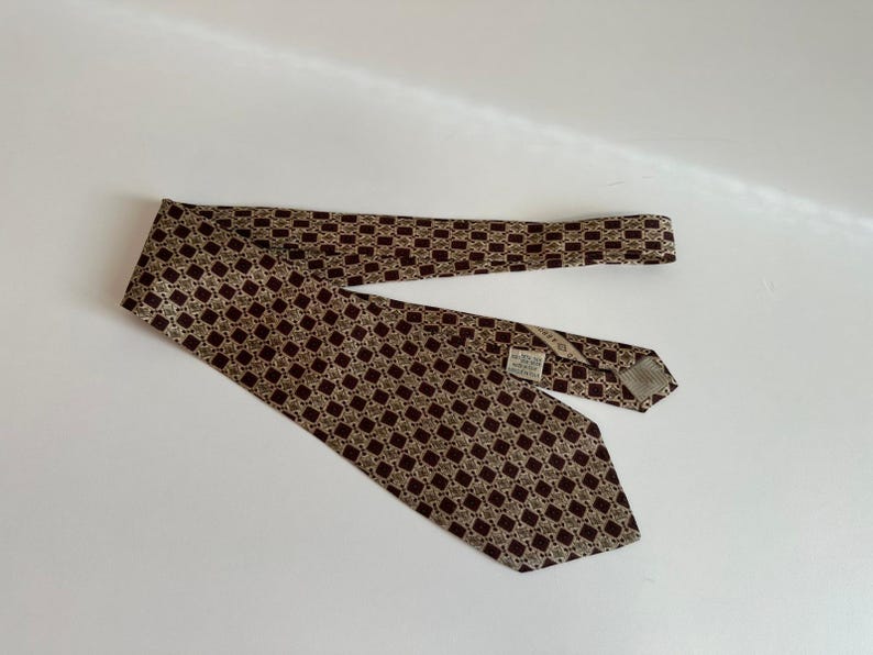 May include: A necktie featuring a repeating geometric pattern of dark brown squares and light beige diamond shapes. The tie has a pointed end and a visible white fabric label stitched onto the back side near the center, displaying printed text.