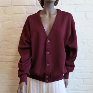 May include: A burgundy cardigan with a V-neck and button closure. The long-sleeved sweater has a ribbed hem and cuffs. The cardigan is displayed on a mannequin in front of a white brick wall.
