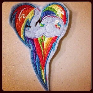 May include: A heart-shaped embroidered patch featuring a rainbow with a blue cloud in the center. The rainbow is made of red, orange, yellow, green, blue, indigo, and violet thread. The cloud has a white outline and a blue fill.