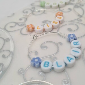 May include: Silver hoop earrings with white letter beads spelling out names like "LISA" and "BLAIR". The earrings also feature small flower-shaped beads in various pastel colors. The background is a light, patterned surface.