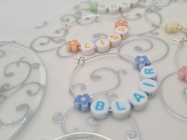 May include: Silver hoop earrings with white letter beads spelling out names like "LISA" and "BLAIR". The earrings also feature small flower-shaped beads in various pastel colors. The background is a light, patterned surface.