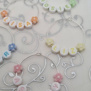 May include: Several hoop earrings with names spelled out in white letter beads. Each earring has a different name, such as "Lisa", "Rosie", "Liz", and "Ashley". The earrings are decorated with small flower-shaped beads in various pastel colors.