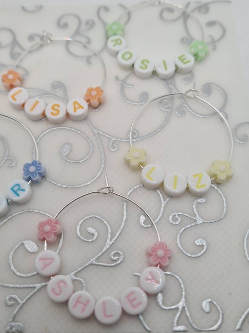 May include: Several hoop earrings with names spelled out in white letter beads. Each earring has a different name, such as "Lisa", "Rosie", "Liz", and "Ashley". The earrings are decorated with small flower-shaped beads in various pastel colors.