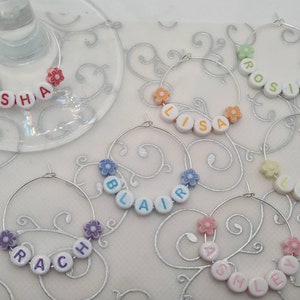 May include: Wine glass charms with names spelled out in white beads with colorful letters. Each charm has a silver hoop and is decorated with flower-shaped beads. Names include Kisha, Lisa, Rosie, Blair, Liz, Rach, and Ashley.
