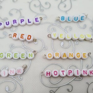 May include: Close-up of several beaded word strands spelling out colors: purple, red, green, blue, yellow, orange, pink, and hot pink. Each word is spelled out with white round beads with colorful letters and silver jump rings.