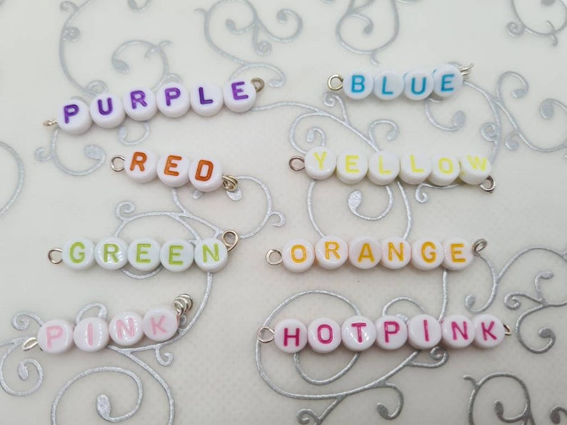 May include: Close-up of several beaded word strands spelling out colors: purple, red, green, blue, yellow, orange, pink, and hot pink. Each word is spelled out with white round beads with colorful letters and silver jump rings.