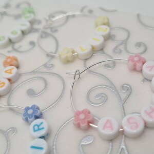 May include: Close-up of several hoop earrings with personalized names spelled out using white letter beads. The beads are accented with colorful flower-shaped beads. The earrings are on a white surface with a silver swirl design.