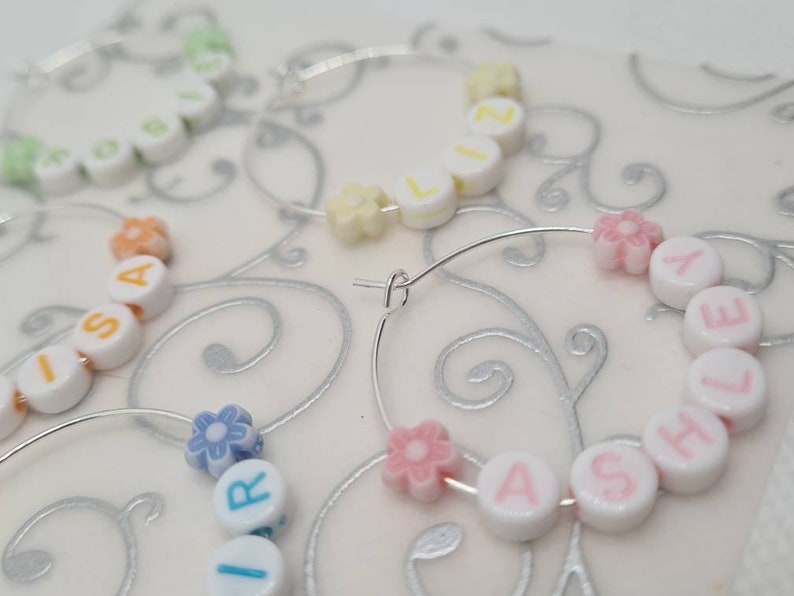 May include: Close-up of several hoop earrings with personalized names spelled out using white letter beads. The beads are accented with colorful flower-shaped beads. The earrings are on a white surface with a silver swirl design.
