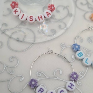 May include: Wine glass charms with names spelled out in white letter beads. The charms are attached to silver hoops and decorated with small flower beads in pink and purple. One charm spells out "KISHA".