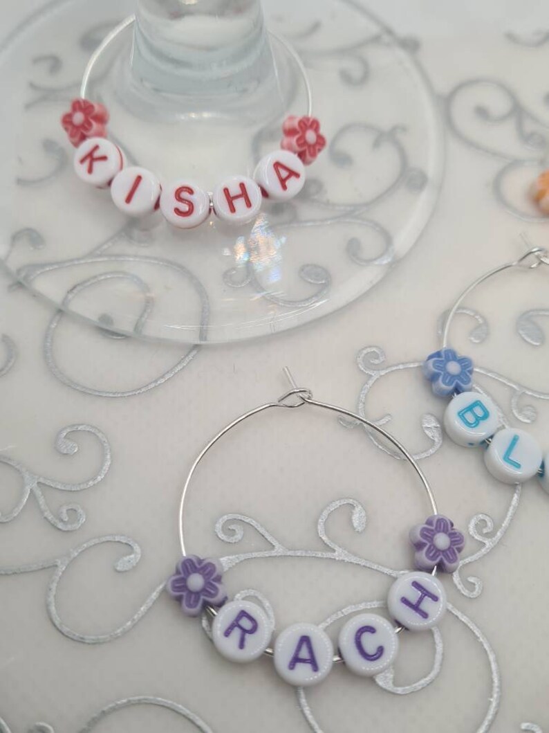 May include: Wine glass charms with names spelled out in white letter beads. The charms are attached to silver hoops and decorated with small flower beads in pink and purple. One charm spells out "KISHA".