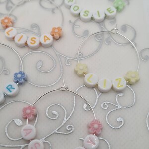 May include: Several silver hoop earrings are displayed, each adorned with white letter beads spelling out names like "LISA" and "ASHLEY." The earrings also feature colorful flower-shaped beads in shades of orange, pink, and yellow.