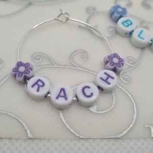 May include: A silver hoop earring with the name "RACH" spelled out in white letter beads with purple letters. The earring is decorated with purple flower beads. Additional beads spell out "BLA".