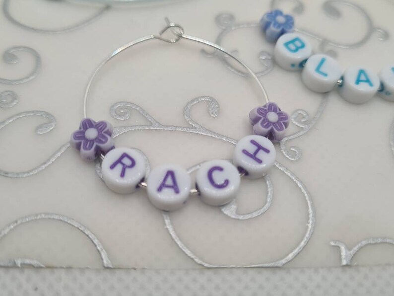 May include: A silver hoop earring with the name "RACH" spelled out in white letter beads with purple letters. The earring is decorated with purple flower beads. Additional beads spell out "BLA".