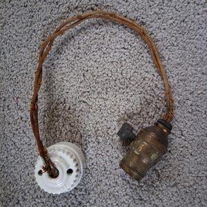 May include: An antique light fixture with a white ceramic base and a brass-colored bulb socket. The fixture is connected by a twisted brown cord. The ceramic base has a decorative edge and two holes.