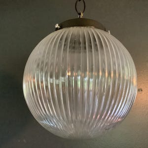 May include: A spherical pendant light fixture featuring a clear, ribbed glass globe. The glass has vertical fluted lines throughout its surface. It is attached to a dark metallic circular cap at the top, which includes a small loop for hanging.
