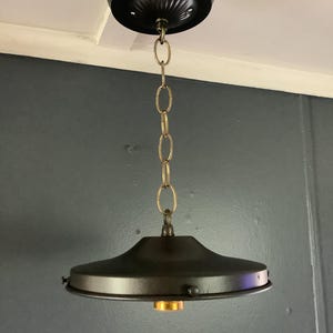 May include: A black pendant light fixture with a circular canopy and a downward-facing, saucer-shaped shade. The shade is suspended by a brass-colored chain. The light fixture is designed for ceiling mounting and has a vintage or industrial aesthetic.