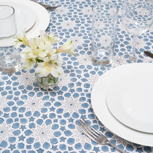 May include: A white tablecloth with a blue and white geometric pattern. There are two white plates, two glasses, a fork, and a small vase with white flowers on the table.