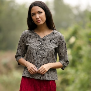 May include: A black and white patterned blouse with a button-up front and three-quarter sleeves. The blouse is worn by a person with long dark hair and is photographed against a blurred green background.