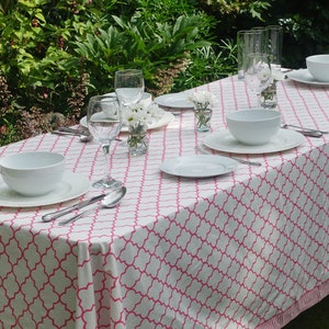 May include: A white tablecloth with a pink geometric pattern is laid out for a meal outdoors. The table is set with white plates, bowls, and cutlery. There are also glasses and flowers in small vases.