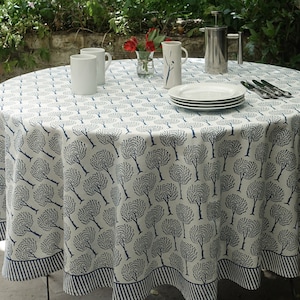 Könnte beinhalten: Eine weiße runde Tischdecke mit einem blauen Baummuster. Die Tischdecke hat einen blau-weiß gestreiften Rand. Die Tischdecke ist mit weißen Tassen, Tellern und Besteck gedeckt.