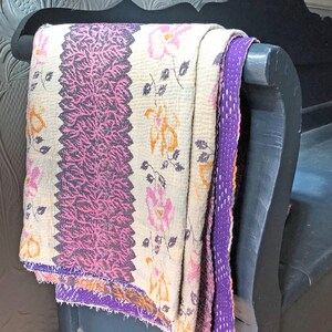 May include: A white kantha quilt with a pink and purple floral pattern. The quilt is folded and draped over a black wooden bench.