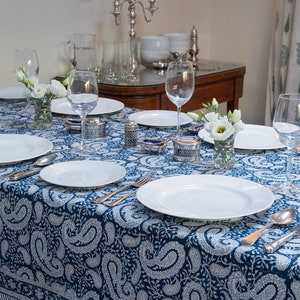 May include: A blue and white paisley patterned tablecloth set for a formal dinner. The table is set with white plates, silverware, and wine glasses. There are small silver containers and white flowers in a glass vase on the table.
