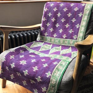 May include: A purple and green kantha throw blanket with a floral pattern. The blanket is draped over a wooden armchair.