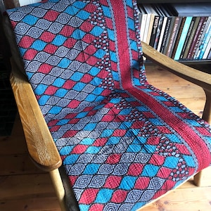 May include: A colorful textile with a geometric pattern of red and turquoise diamonds and circles, draped over a wooden chair. The fabric has a quilted appearance and a red border. The chair is made of light-colored wood.