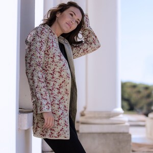 Könnte beinhalten: Eine Frau trägt eine langärmelige, beige Jacke mit rotem Blumenmuster. Die Jacke hat einen Stehkragen und eine lockere Passform. Sie steht vor einer weißen Wand.