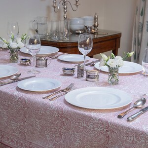 May include: A pink and white patterned tablecloth with white plates, cutlery, and wine glasses set for a formal dinner. White flowers are in a small glass vase in the centre of the table.