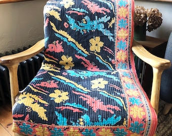 May include: A black and blue floral patterned throw blanket with a pink and yellow border. The blanket is draped over a wooden chair.