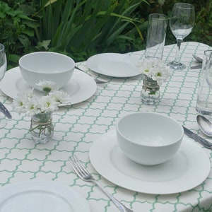 May include: A white tablecloth with a green trellis pattern, set for a dinner party. There are two white bowls, two white plates, two glasses of water, two wine glasses, two forks, two spoons, and two knives. There are also two small vases with white flowers.