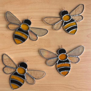 May include: Four stained glass bee ornaments. Each bee has a black and yellow striped body, clear wings, and a black head. The bees are displayed on a light brown wooden surface.