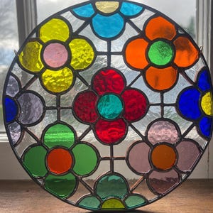 May include: A round stained glass window with a floral design. The flowers are in shades of yellow, pink, orange, green, red, and blue. The glass is set in a black metal frame. The window is hanging in front of a window.