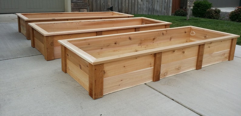 May include: Three wooden raised garden beds made of cedar wood. The beds are in a row, with the largest bed in the foreground and the smallest bed in the background.