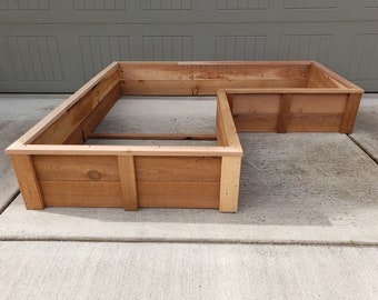 May include: A wooden raised garden bed in the shape of an L. The bed is made of natural cedar wood and is empty.