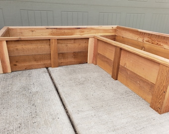 May include: A corner wooden planter box made of cedar wood. The planter is made of multiple pieces of wood that are joined together. The planter is sitting on a concrete surface.