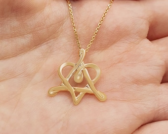 Puede incluir: Un collar de oro con un colgante en forma de corazón formado por la letra A. El colgante está en una cadena y se sostiene en una mano.