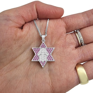 May include: A silver pendant necklace with a pink and white Hamsa hand charm inside a Star of David. The charm is set with small pink and white stones.