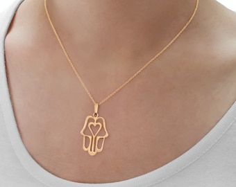 Puede incluir: Un collar de oro con un colgante de mano de hamsa con un corazón recortado. El colgante es un diseño delicado y calado.