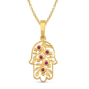 Puede incluir: Un collar con colgante dorado con un diseño de mano de Hamsa. El colgante está adornado con piedras preciosas rojas y un motivo floral. El collar tiene una cadena delicada.