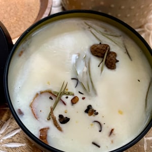 May include: A white candle with a black rim and a green lid. The candle is filled with white wax and has various natural elements embedded in it, including dried herbs, pine cones, and spices.