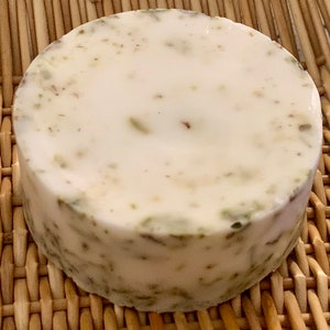 May include: A round, white soap bar with green flecks throughout. The soap is sitting on a woven wicker surface.